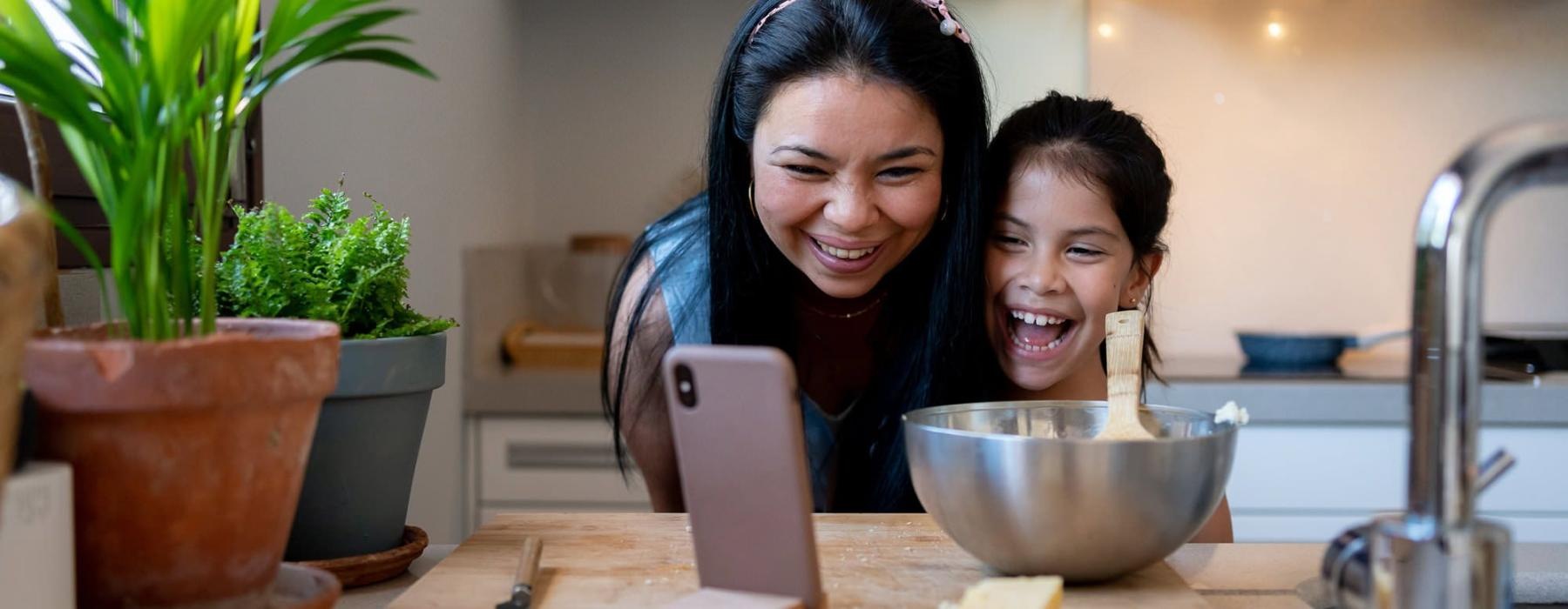 mother and young daughter smile at their phone as they cook in the kitchen